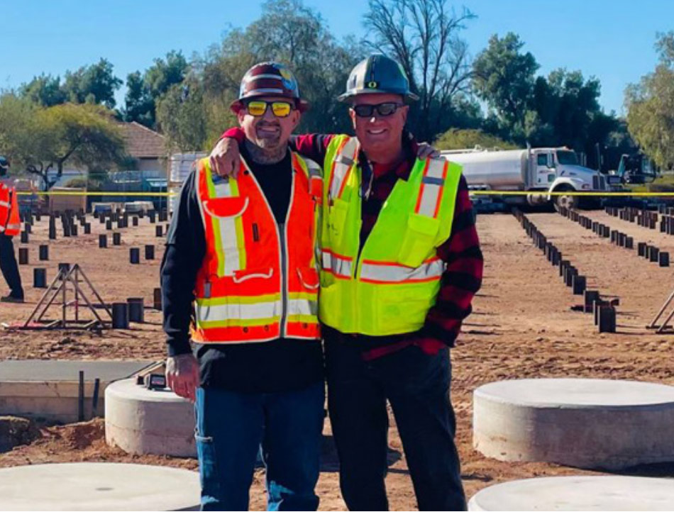 two men in hard hats and contractor vests with an arm around each other