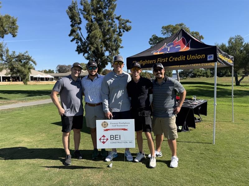 A group of five BEI Construction employees standing on a golf course behind a sponsorship sign for the Fremont Firefighters Golf Tournament, with an event canopy in the background.
